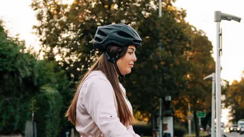 A woman wearing a cycle helmet and light pink jacket parks up her ebike on a street station along a residential road.