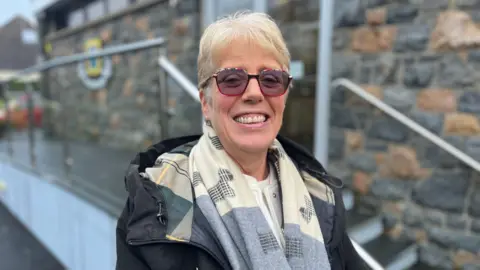 A woman standing outdoor is smiling at the camera. A stone wall and steps are behind her. She is wearing sunglasses and a scarf. 
