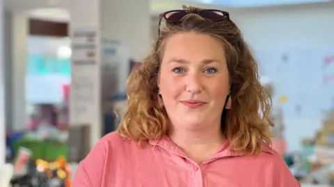 A woman with shoulder-length, wavy blonde hair. She has sunglasses on top of her head and wears a pink blouse and pink tassled earrings. Blurred images of food stacked on tables can be seen behind her.