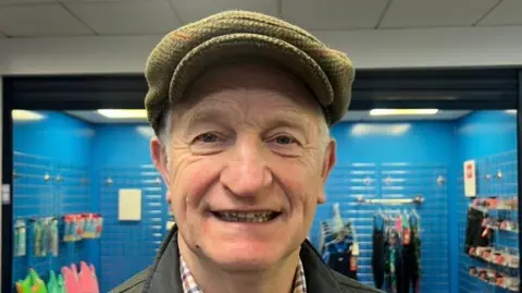 Paddy Cosgrove is wearing a green hat and jacket. He has short grey hair and is standing near the swimming pool's shop.
