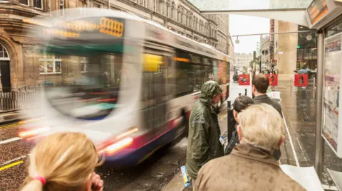 A bus arriving at a Glasgow bus shelter on a rainy day, with several people waiting, with their hoods up