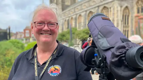Alex Dunlop/BBC Bev Coleman is wearing a black polo shirt, branded with Cromer Peregrine Project. She is also wearing a black branded lanyard and is standing next to a camera pointed at the top of Cromer church.