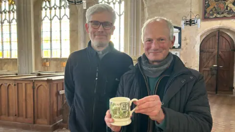 Immy Lee/BBC Jonathan Dowman has white short hair, swept back and is wearing a navy fleece and a blue shirt. He is smiling and standing next to Charles Killin, who has grey hair and is wearing a grey scarf, jumper, navy fleece and black padded coat. He is holding the mug and smiling.