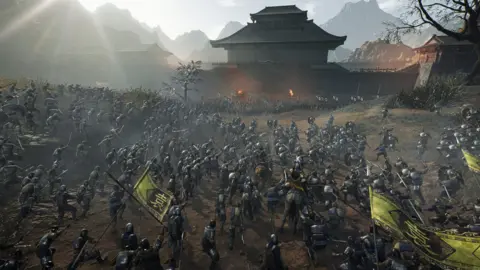 Koei Tecmo A battle sequence from the game. There are many hundreds of soldiers in feudal era armour running towards one another holding pikes, swords and other similar era-appropriate weapons. There is a Chinese temple in the background, with mountains behind.