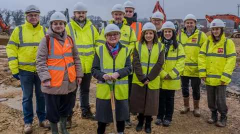 FIona Baker with glasses and a white hard hat with a yellow hi-vis jacket holding a spade, standing on waterlogged soil with 10 other people similarly dressed, although one has an orange hi-vis. There are trees behind.