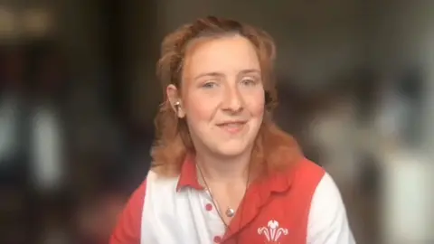 Lillith is wearing a red and white striped Wales rugby shirt. She has medium length red hair.