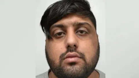 West Yorkshire Police A man with black hair and a black beard looks towards the camera. He is wearing a grey top and stands against a plain white background.