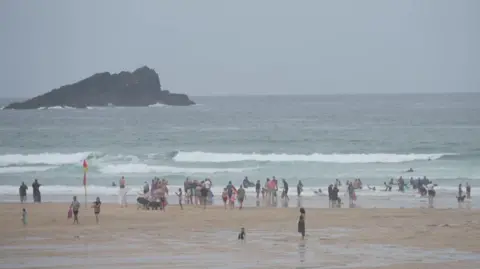 There are lots of surfers and people on the beach at Fistral Beach in Newquay. There is a large rock sticking out of the water and large waves smashing against the beach. 