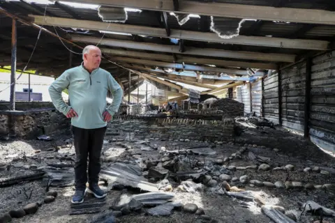 Lorcan Doherty A man wearing a blue jumper stands in the wreckage of a fire. he has his hands on his hips and stares into the devastation. All around his are charred remains of what the museum once was.
