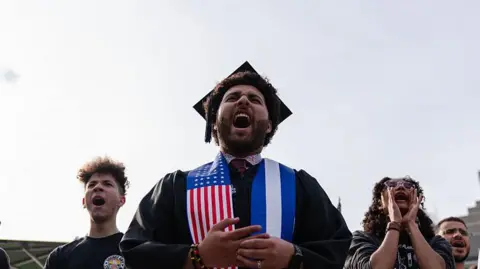 A Harvard University student chants with fellow demonstrators during a protest in support of international students at Harvard University.  He is wearing a graduation cap and gown with a sash which has an American flag on it. 
