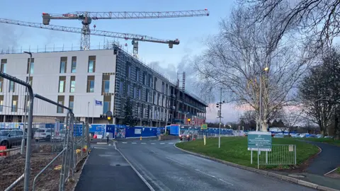 Building work at Royal Shrewsbury Hospital. Two cranes tower over a four-storey building, with metal fences on the left and a car park on the right