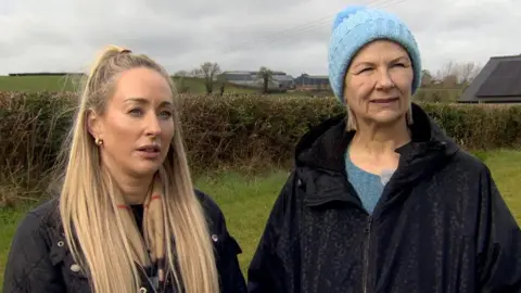 Two women in coats standing beside one another outside in front of a field looking at the camera. The sky is grey, the field is muddy and there are flowers behind them.