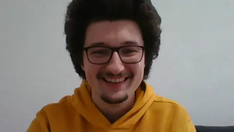 Frank Ilett smiling into a webcam camera. He is wearing a mustard coloured hoodie, black glasses, and has raised long dark hair.