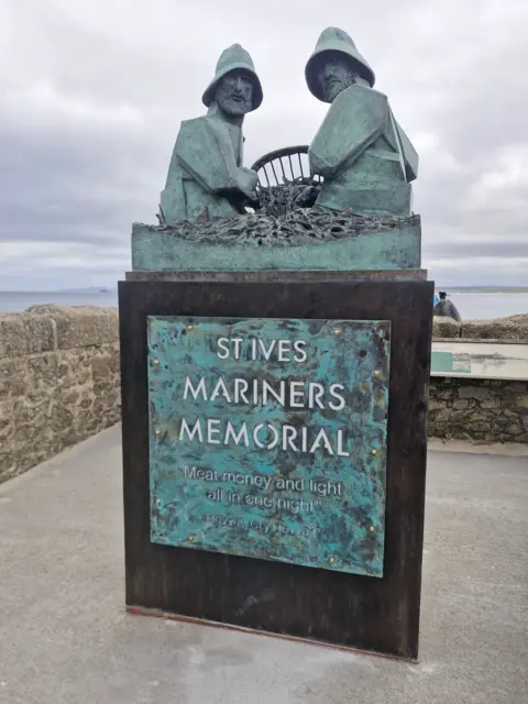 Tom Leaper Bronze St Ives Mariners Memorial sculpture of two fishermen in oilskins beside a ship's wheel, overlooking the sea under a cloudy sky.