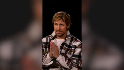 Ryan Gosling presses his hands together. He is wearing a white fleecy jacket, with large black and orange check lines.