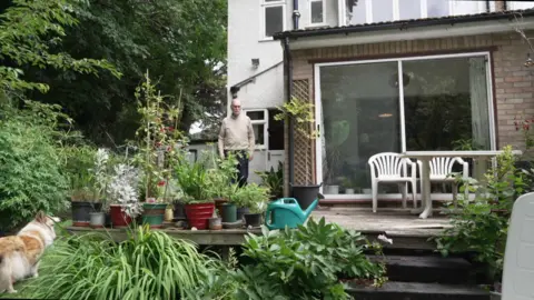 Jim in a beige jumper stands on a wooden decking area outside a house, surrounded by potted plants and greenery, with a dog in the foreground