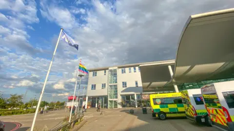 Darent Valley Hospital in Dartford, England, with people visible in the background and the NHS and Gay Pride flags outside.