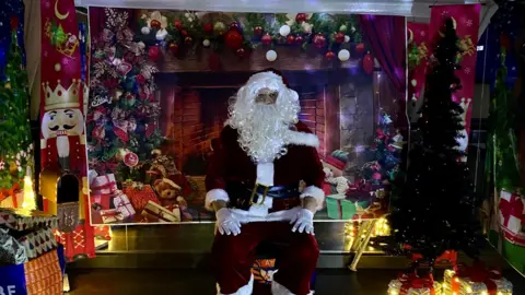 A Santa surrounded by Christmas presents and a decorative Christmas backdrop.