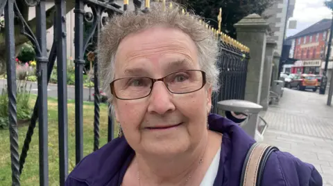 Sandra McElheron tiene el pelo rizado gris/castaño claro y gafas de montura negra, y lleva un abrigo morado.