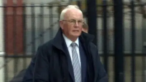 Frank Daly is wearing a black coat over a navy blazer, white shirt and stripped tie. He has glasses on and short grey hair on his head. There is a person behind him. The background is blurry. 
