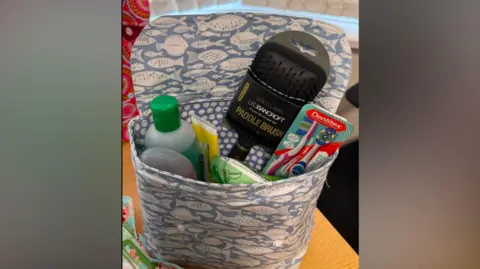 The Haven One bag is blue with a fish patter and has several toiletries inside it. It is laid on a table,