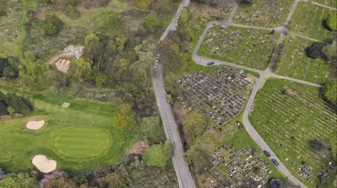 An ariel shot showing a carriageway, parked cars on a field and golf course.