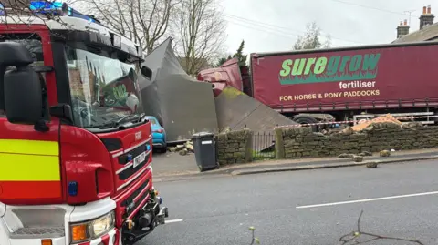 A red lorry which has crashed through a bricked wall and then into an outbuilding. The lorry has the words 'Suregrow fertiliser for horse and pony paddocks' written on the side. In the foreground is a red fire engine parked on the road side.