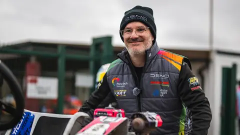 Josh East Photography Chris looks and smiles at a brightly coloured kart. he wears glasses, a sponsored gilet and a beanie