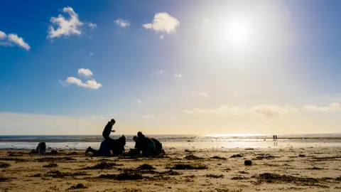 Dru Dodd A group of children are building a sandcastle on a beach with the sea behind them. The sun is low in the sky so they are mostly black shapes with a stretch of sand behind lit by the suns rays. 