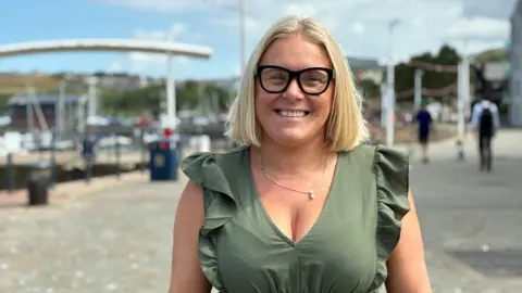 Emma Williamson has blonde hair and glasses, is wearing a pale green dress and is looking straight ahead, standing on Whitehaven Harbour