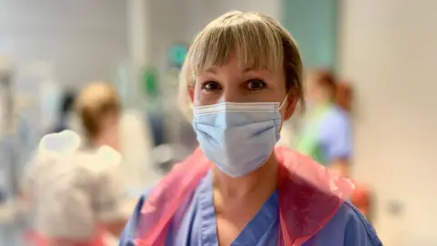 Mark Norman/BBC ICU senior sister Lucy Gosnell poses for the camera wearing a mask and apron with patients and colleagues behind her.