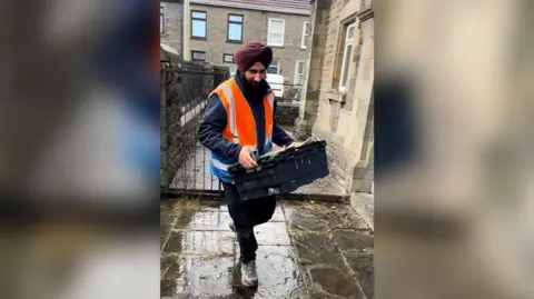 Marcus Lapsa Bill Mato carries a box of aid into the stone-built Trallwn Community Centre in Pontypridd. Terraced houses can be seen behind him and there is water on the pavement.