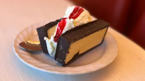 Two scoops of ice cream covered in sauce are sandwiched between chocolate covered, mallow-filled wafers. The treat sits on a white plate on a table.