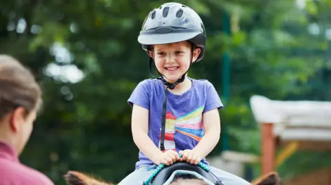 A young child smiles as they sit on a horse and hold on to a blue rope. An instructor is in the foreground of the photo.