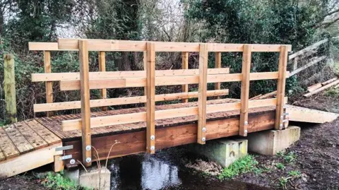 Butterfly Conservation A completed wooden footbridge spans a small stream in a natural, wooded area. 