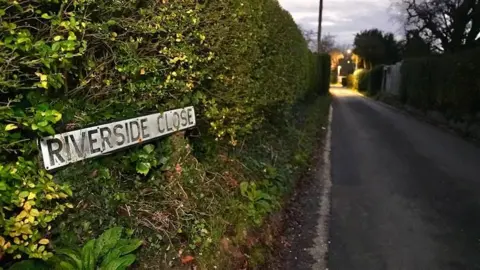 Martin Giles/BBC A road sign saying "Riverside Close". It is in a thick green, hedge that stretches back into the distance along a single-track road. A light, possibly a street light, can be seen in the distance. The picture has been taken at dusk.