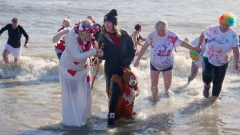 People in the North Sea at Felixstowe wearing fancy dress. They are exiting the water after taking part in a Valentine's Day dip on a sunny day. Two are holding hands.