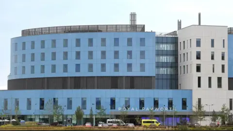 PA Media The exterior of Royal Papworth Hospital. A large hospital with several floors. Much of the modern building has blue cladding. The words "Royal Papworth" are in big letters over a covered entrance. There are vehicles, including an ambulance, in front of the hospital.