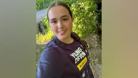 Jasmine Barker standing on the side of a road, surrounded by greenery and flowers, smiling to the camera and wearing a navy blue running top with Young Devon Run Devon written on the front