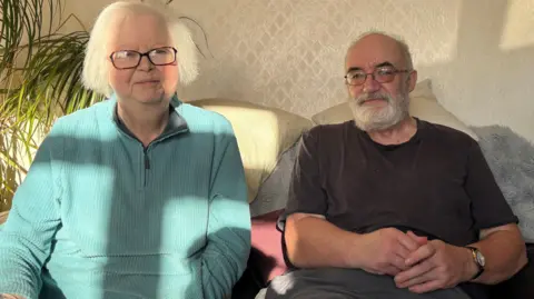 A woman in a light blue jumper sits next to her husband who is wearing a grey shirt, on a sofa. Behind them on one side are green fronds of a plant and behind them is beige patterned wallpaper