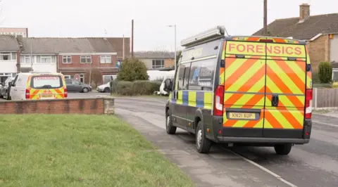 A police forensics van parked on the street, partly on the pavement. Another smaller van is parked on a driveway. It is a residential street with a number of terraced and semi-detached homes visible in the background. 