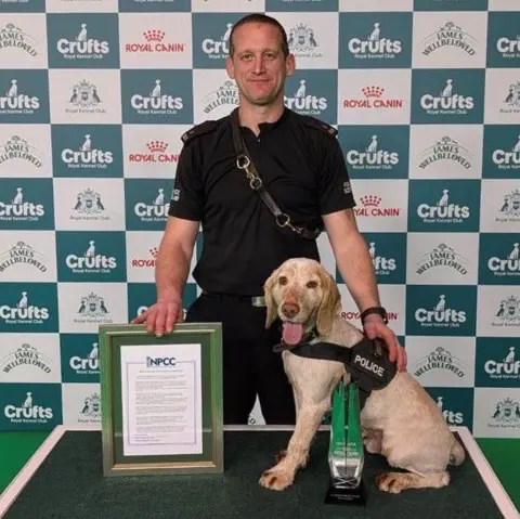 Police Scotland Mac and his handler Andy Inglis. Mac is on a podium and Andy is standing beside him with a framed certificate