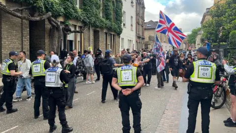 A row of police officers stand in the street in front of a group of people holding union flags.