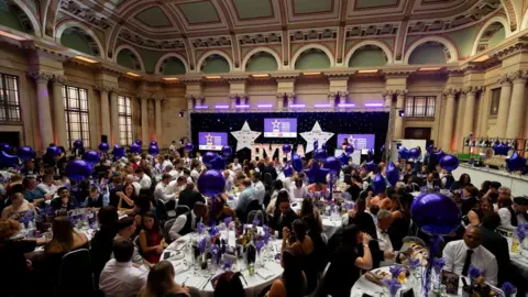 Dougie Allward/Made For Impact A wide shot of the room where the awards ceremony took place. There are many round tables full of people and lots of purple balloons. The stage can be seen at the opposite end of the room.
