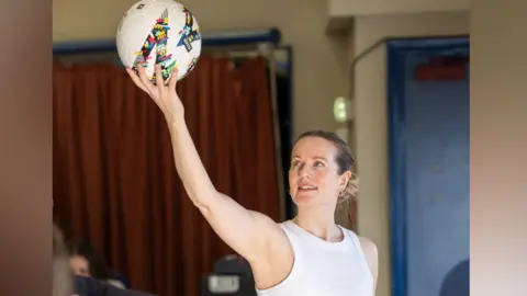 A woman wearing a white vest top holds a football above her head