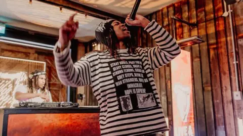 A man in a black and white striped shirt hold a microphone. He has long black hair and has on a beanie. Behind him is a DJ spinning turntables