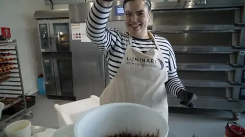 A woman with a striped top sprinkles powder into a bowl of cake mix, wearing an apron saying Luminary Women Bakery