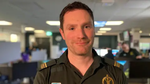 Karl Bloomer - a man wearing a green ambulance uniform with gold lapel details and a logo smiles at the camera. He is standing in a call centre, but the background is blurred.