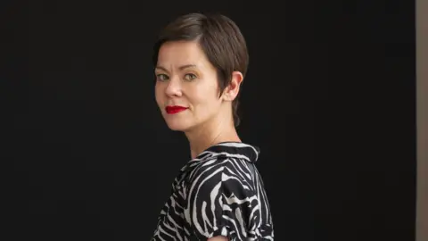 Christopher Owens Laura Brewis smiling and  looking over her shoulder. She has a short, straight brown hair and wears a midi length zebra print dress with short sleeves.