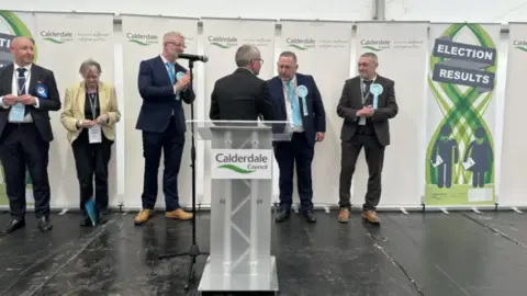 A line of political party candidates stand on a podium, in front of signs reading Calderdale Council. 